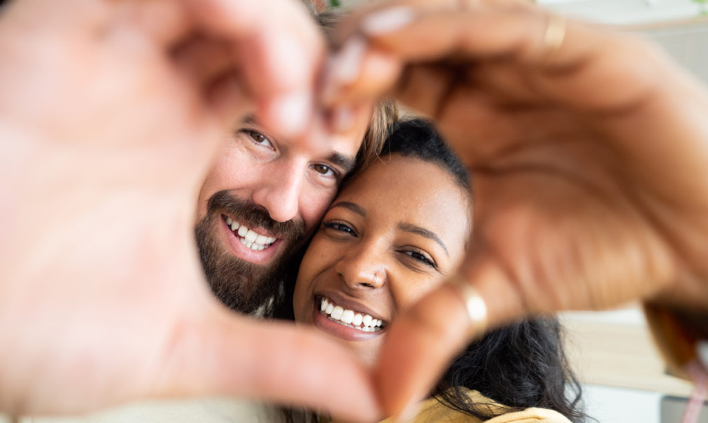 Smiling couple at home making a heart shape with their hands in the foreground