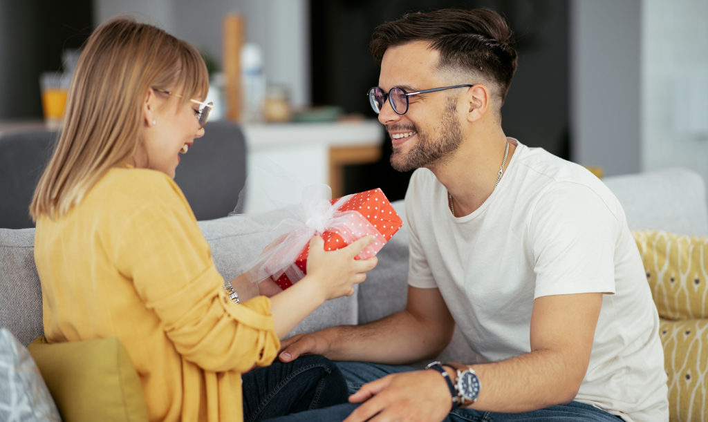 Husband surprising his wife with a gift while they are seated on their living room couch