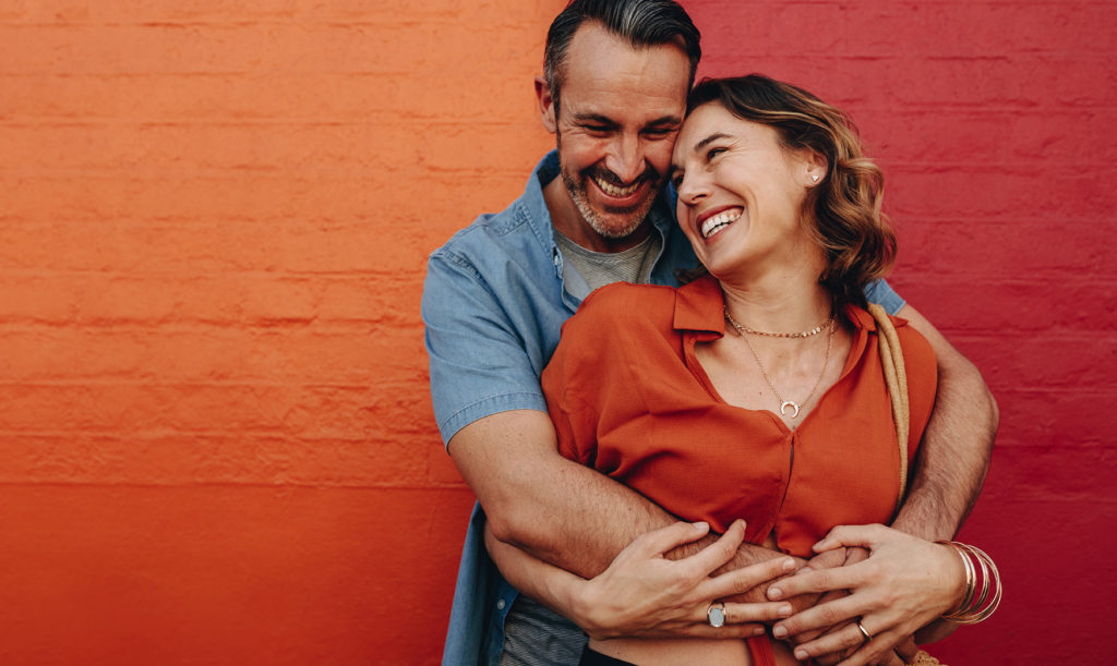 Married couple smiling and embracing standing against a wall