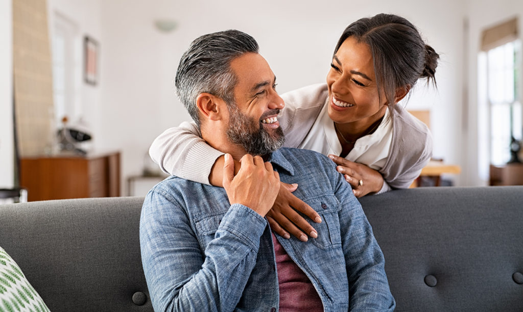 Smiling woman hugging her husband on the couch from behind in the living room