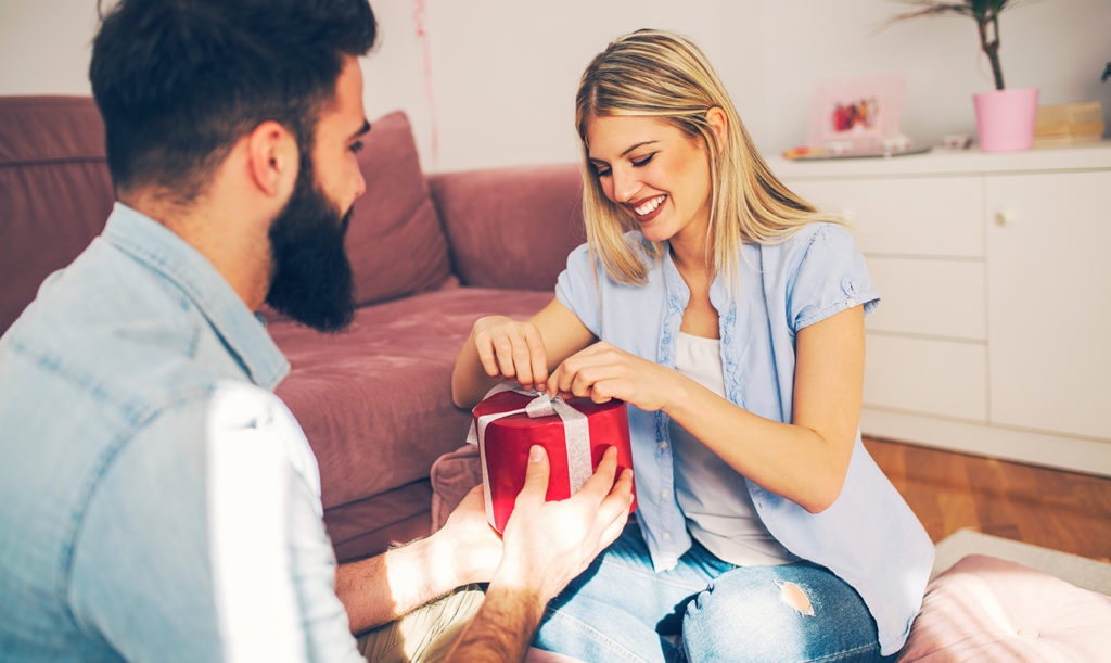 Happy woman opening a gift from her husband seated on the floor of their living room