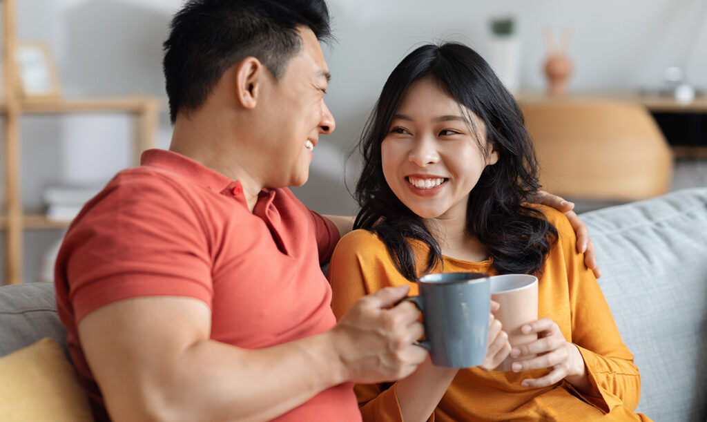 Casual photo of happy loving asian couple sitting on couch in living room, drinking coffee from ceramic mugs while smiling at each other