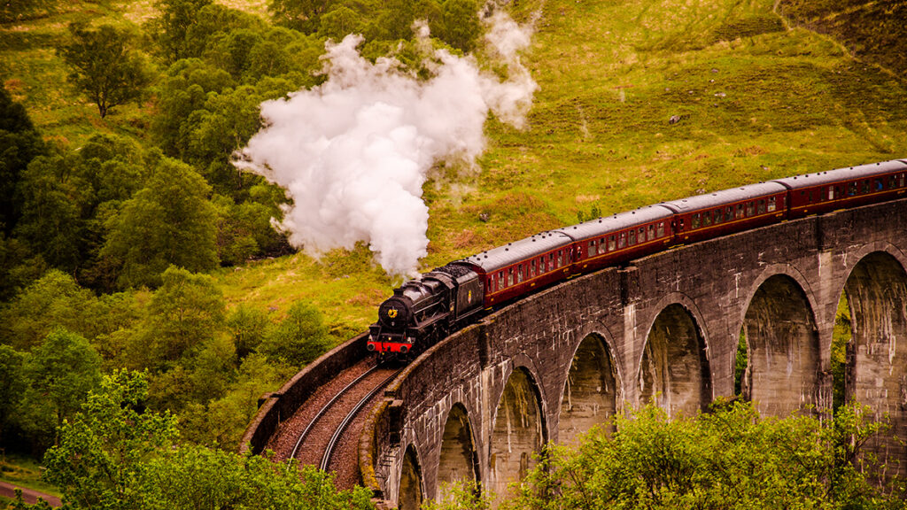 Image of a train resembling the Hogwarts Express travelling over the Glenfinnan Viaduct in northern Scotland with steam billowing from the engine car surrounded by green hills.