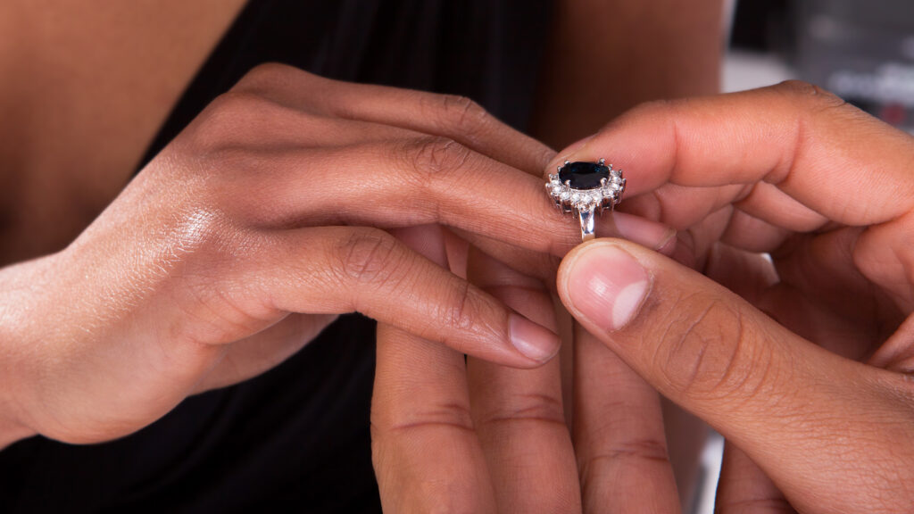 Close up of a male hand placing a sapphire and diamond halo ring on the ring finger of his wife's right hand