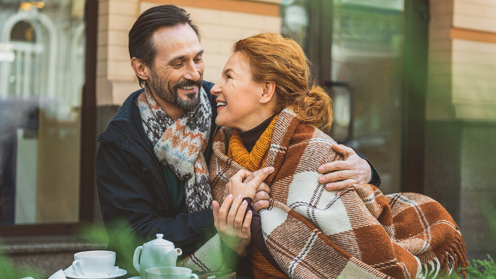Image of a middle aged white couple seated at an outdoor table cuddling in a cozy blanket and scarves while drinking tea