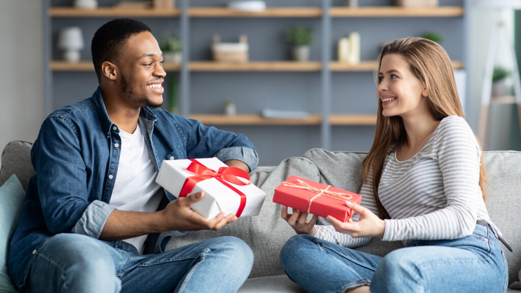 Candid image of interracial couple exchanging wrapped anniverasry gifts while sitting on their couch at home