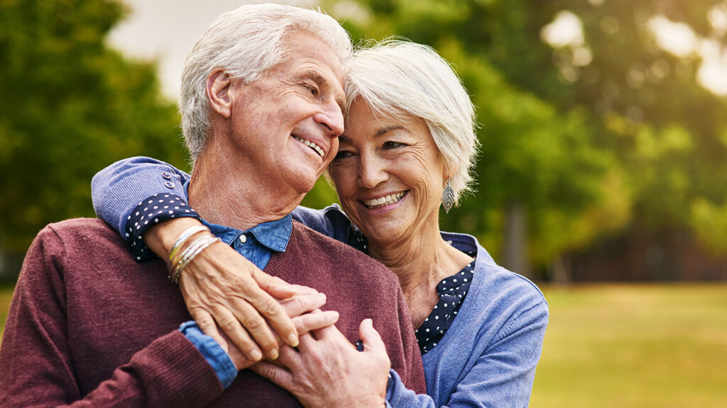 Candid image of a older mixed race couple smiling in a park outdoors with the wife standing slightly behind to husband giving him a loving embrace.