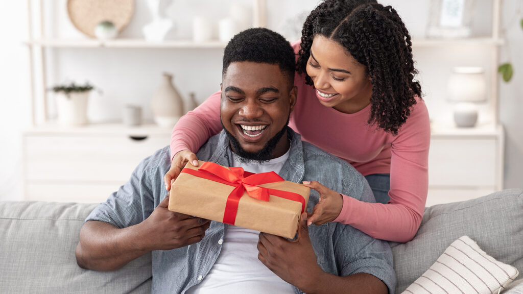 Candid image of a smiling Black couple at home with the husband seated on the couch and the wife leaning over the back, surprising him with a wrapped anniversary gift.