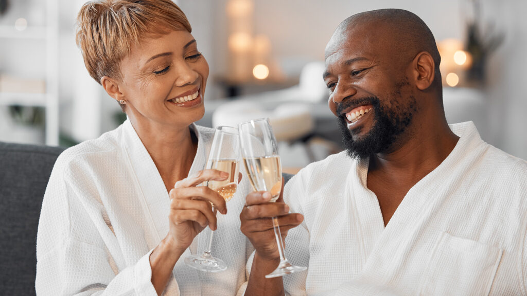 Candid image of a Black couple, celebrating their anniversary at home in spa robes while smiling and toasting with champagne