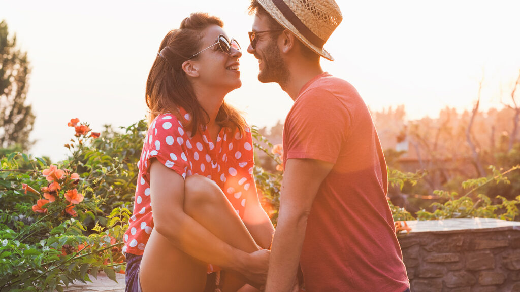 Candid image of a couple outdoors during sunset with the wife sitting on a rock wall wearing a red and white polks dot shirt with her husband leaning in to kiss her 