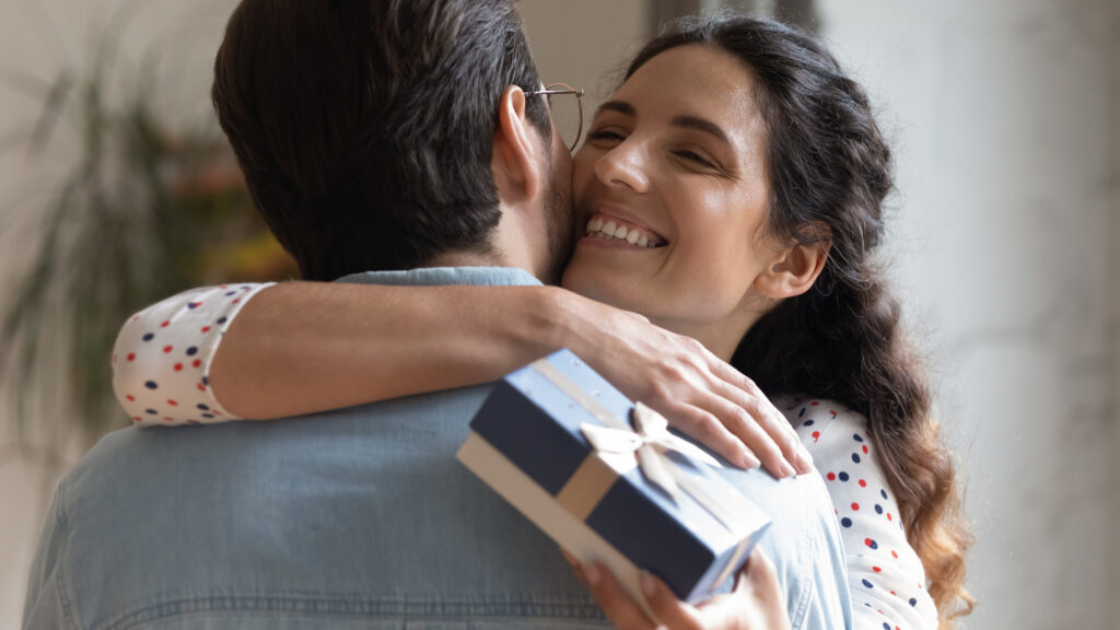 Close up overjoyed wife holding gift box with bow while hugging her husband in thanks for the romantic anniversary present