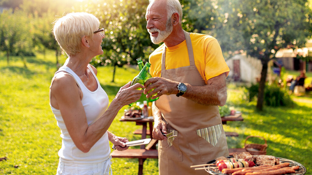 Candid image of an older couple cheersing with beers while grilling outdoors