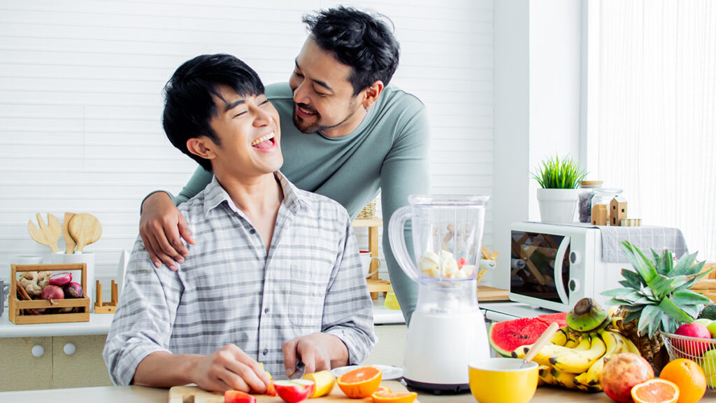 Candid image of a gay asian couple at home in their kitchen laughing with each other while preparing a fruit smoothie.