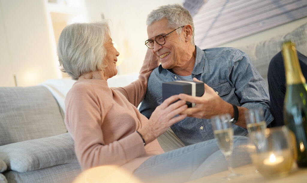 Featured image for 40th anniversary gift guide - a candid photo of a senior couple smiling at each other while celebrating their anniversary by exchanging gifts at home.