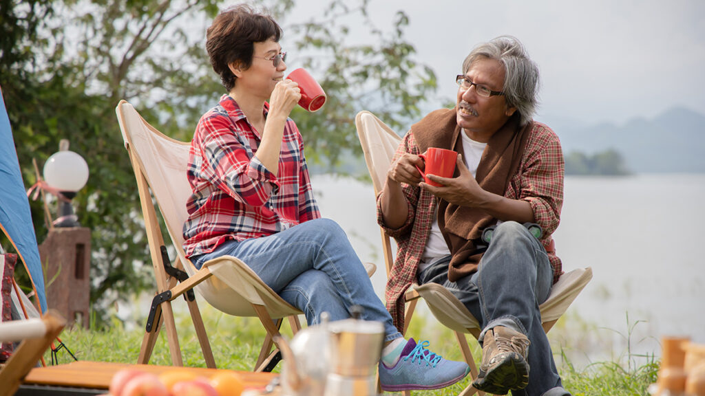 Happy retired couple seated in canvas camp chairs having coffee together by their tent