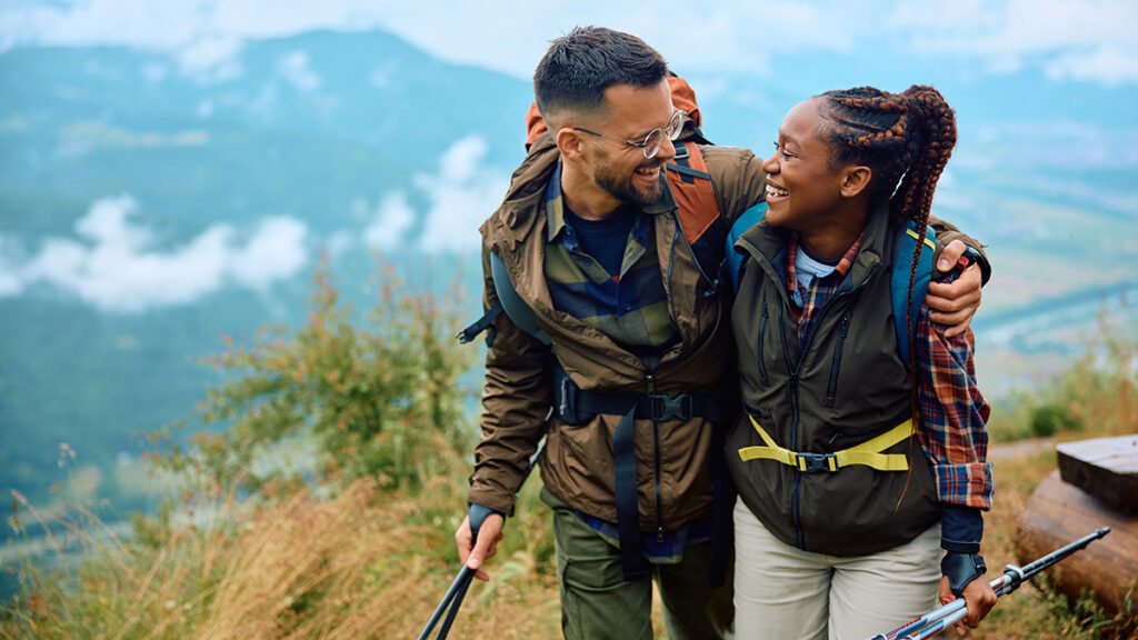 Multiracial couple embracing and smiling at each other while hiking on a mountain with backpacking gear.