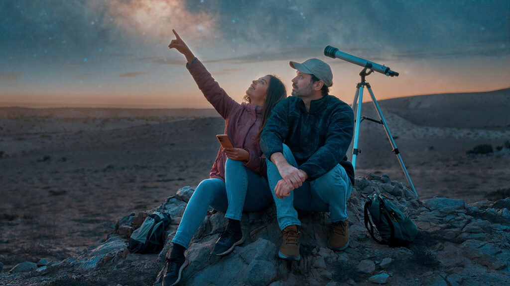 Young couple sitting in the desert stargazing next to a telescope with the Milky Way in the sky.