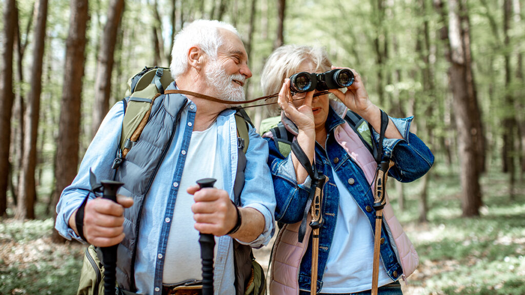 Senior couple with hiking backpacks and trekking sticks in the forest looking at wildlife through binoculars